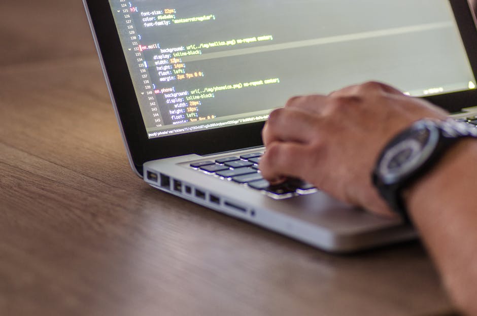 A close-up image of a person's hand operating a laptop computer on a wooden surface. The laptop screen displays lines of HTML or CSS code, indicating coding work or web development. The person is wear