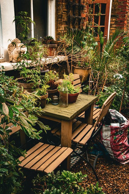 A small urban courtyard garden situated against a brick wall with a black framed window. The space features a white metal bistro table and matching chairs, with one chair partially seen in the foregro